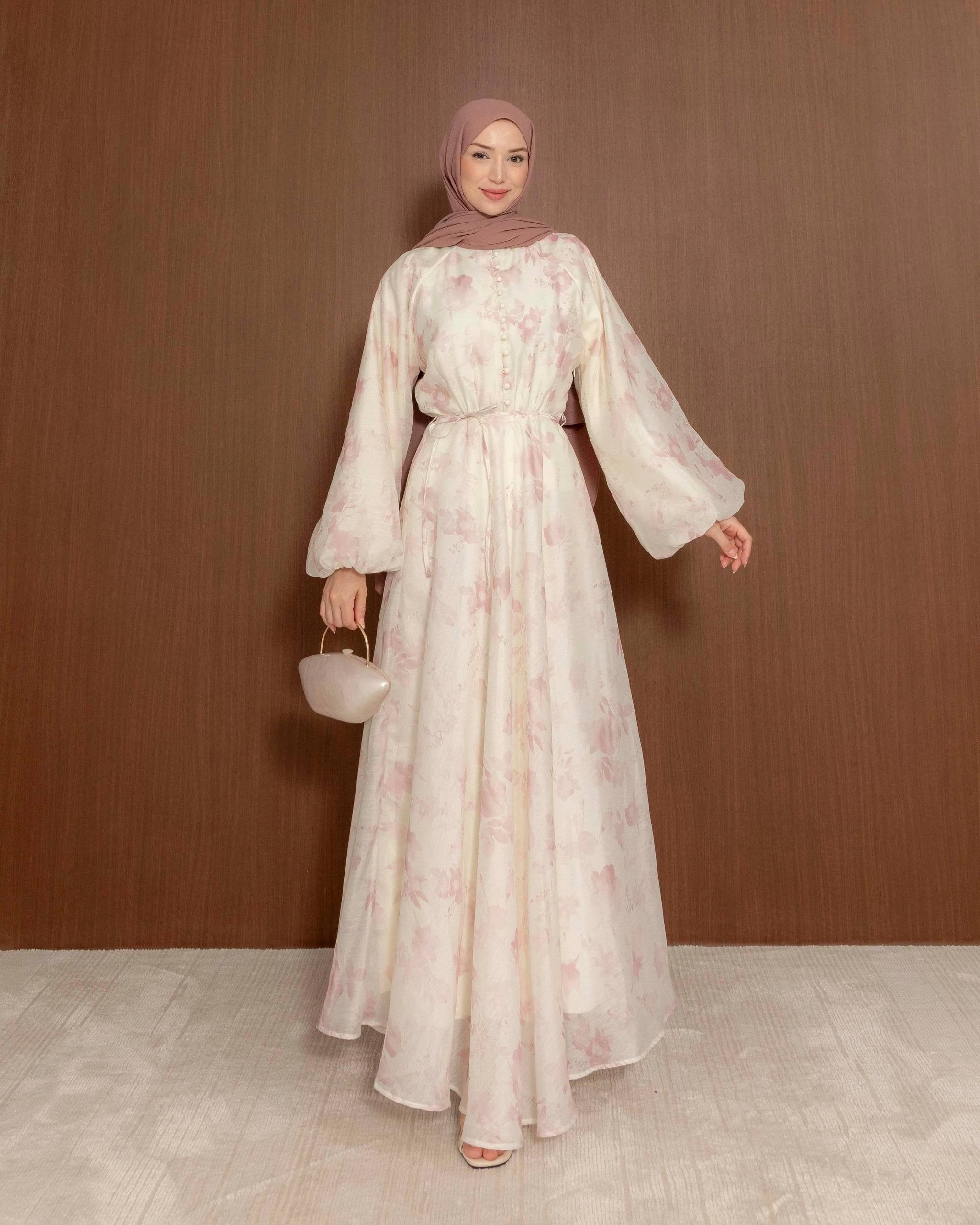 A woman wearing a long, flowing cream-colored modest dress with a light pink floral pattern, a matching pink hijab, and holding a small cream handbag, standing against a brown wooden wall.
