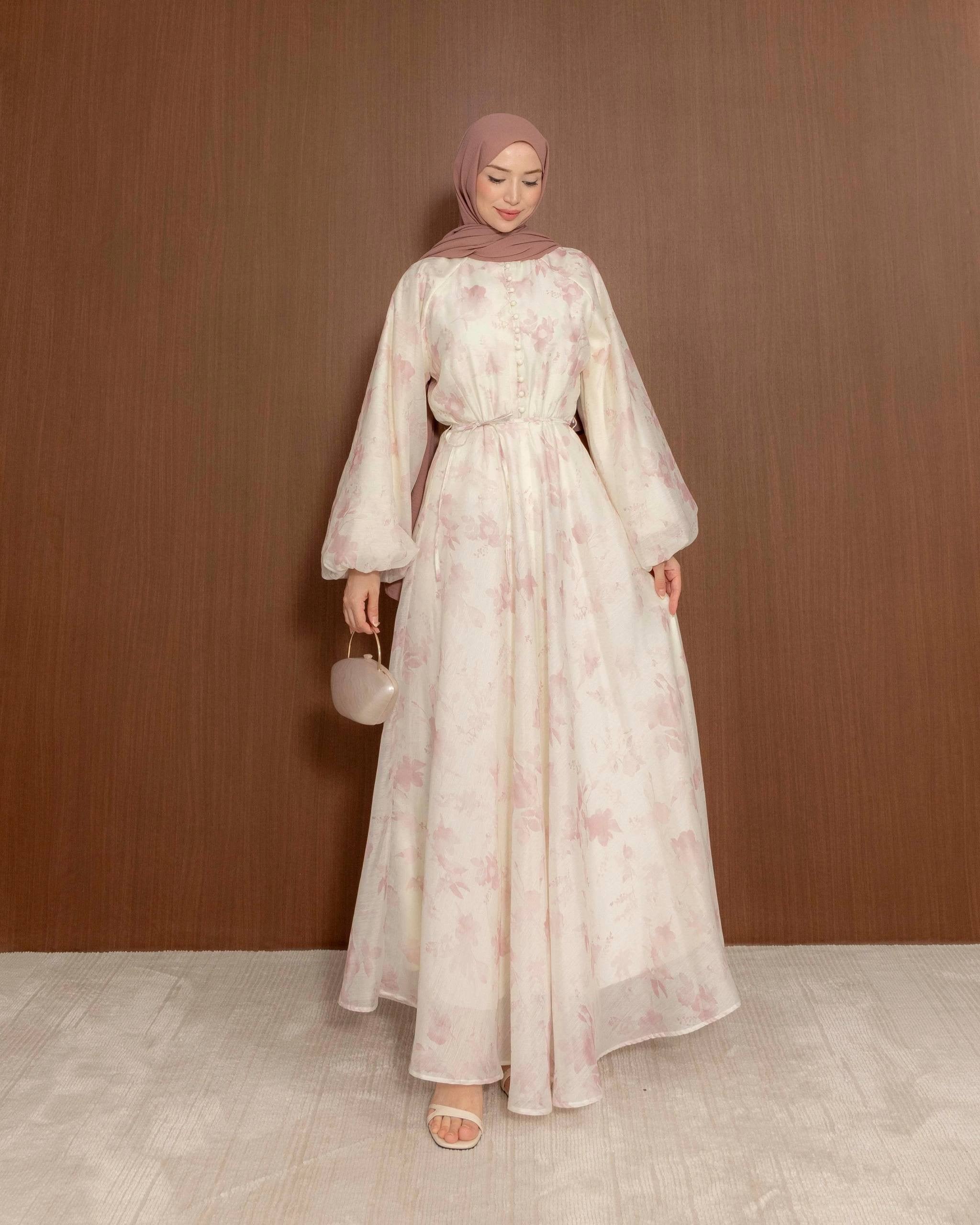 A woman wearing a long, flowing modest dress in a soft off-white with a light pink floral pattern, paired with a dusty rose hijab and holding a small matching handbag, standing against a brown wooden wall.