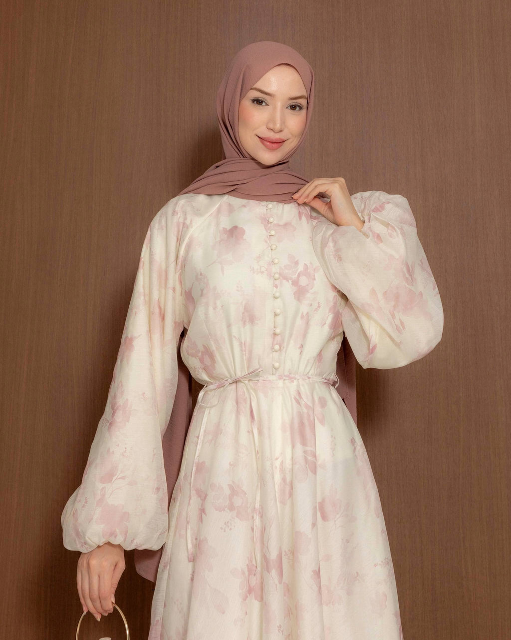 A woman wearing a long-sleeved, button-front modest dress in a light cream color with a soft pastel pink floral pattern, paired with a dusty rose hijab, standing against a brown textured background.