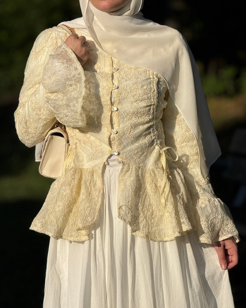 A woman wearing a cream and ivory traditional outfit with a lace-embroidered asymmetrical jacket, matching headscarf, and long skirt, holding a small cream handbag in warm outdoor light.