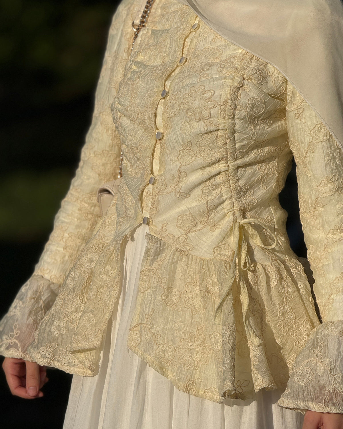 A close-up of a person wearing a cream-colored lace and satin historical-style dress with a ruffled bodice, long sleeves, and a white skirt, photographed outdoors in warm light.