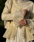 A close-up of a person wearing a cream and ivory embroidered lace jacket with a matching sheer headscarf and white skirt, holding a small cream handbag.