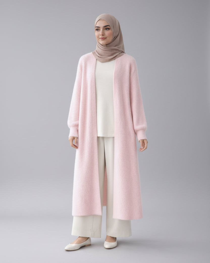 Woman wearing a light pink cardigan, white top, and pants with a beige hijab in a minimalistic setting.