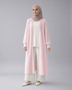 Woman wearing a light pink cardigan, white top, and pants with a beige hijab in a minimalistic setting.