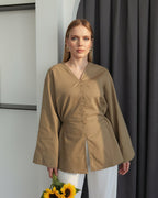 A woman stands indoors wearing a loose, long-sleeve taupe-brown top with a V-neck and front buttons, paired with white pants. She holds a small woven handbag with a yellow sunflower accent.