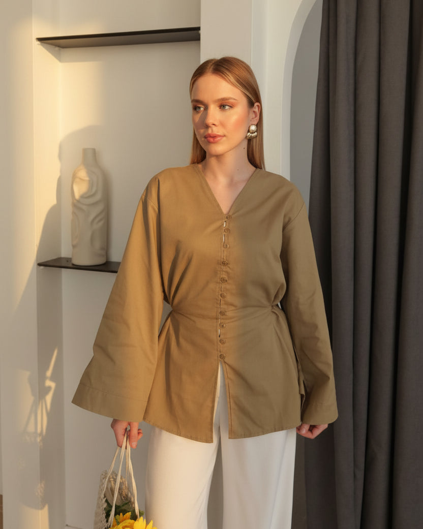 A woman models a taupe-brown long-sleeve button-up top with a V-neck and front slit, paired with white pants and holding a small woven tote bag with yellow flowers, set against an indoor background with white walls, black shelves, and a dark gray curtain.