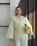 A woman wearing a pale yellow long-sleeve button-up blouse and white pants, holding a small woven basket with yellow flowers, standing in front of a dark gray curtain and white wall.