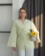 A woman wearing a pale yellow long-sleeve button-up blouse and white pants, holding a small woven basket with yellow flowers, standing in front of a dark gray curtain and white wall.