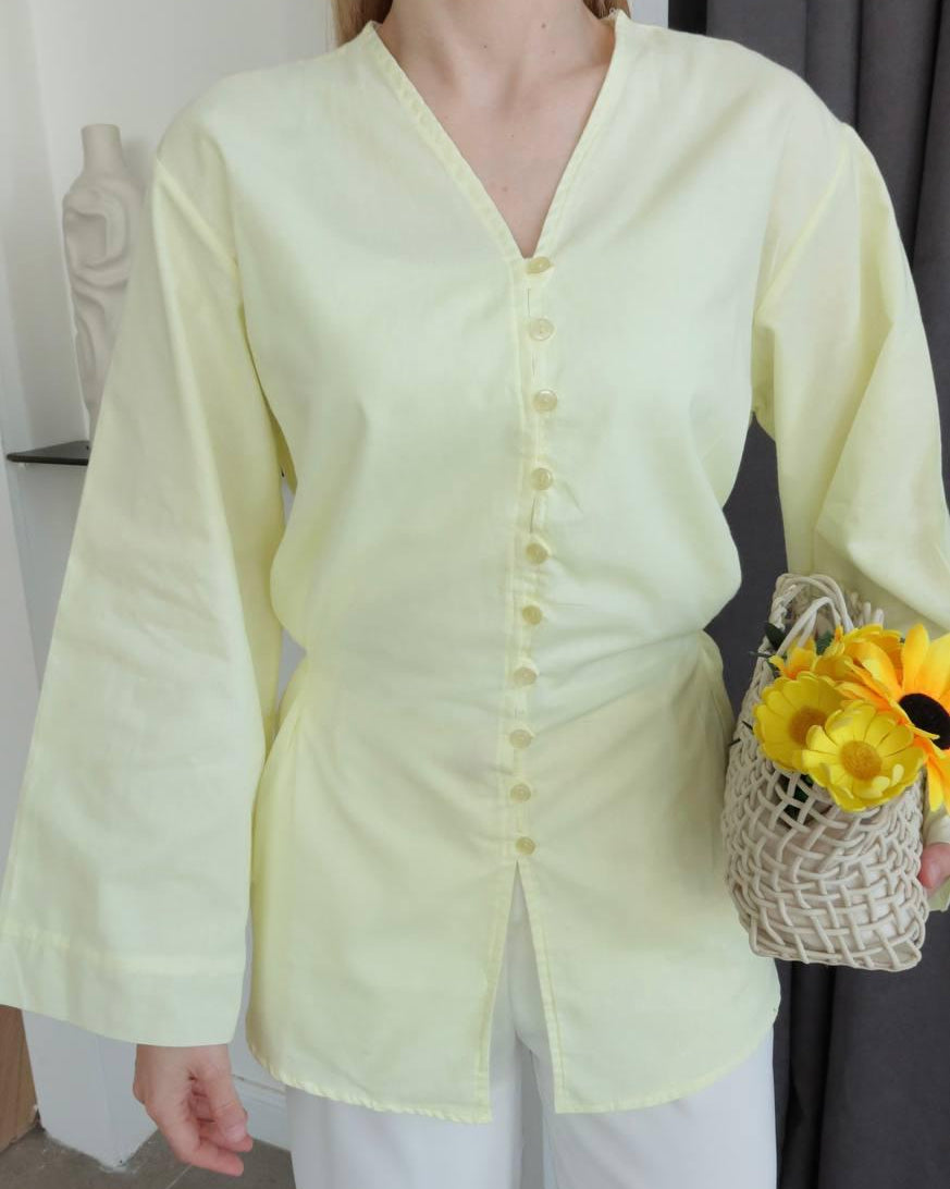 A model wearing a pale yellow long-sleeve button-front top with a V-neck and white pants, holding a woven basket of yellow flowers in an indoor setting.