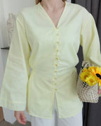 A model wearing a pale yellow long-sleeve button-front top with a V-neck and white pants, holding a woven basket of yellow flowers in an indoor setting.