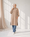 Woman wearing a beige long-sleeve top and blue jeans in a bright room.