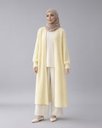 Woman wearing a long light yellow cardigan over a white outfit with a beige hijab on a white background